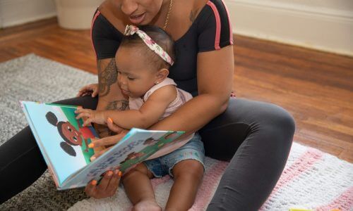 A photo of a woman sitting on the floor holding a baby and reading book