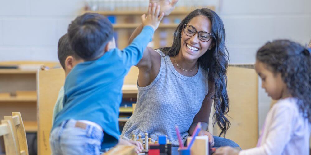 A female Montessori teacher sits with a small group of students as she works through a math lesson. They are each dressed casually and listening attentively as the teacher holds up the numbers in front of her.