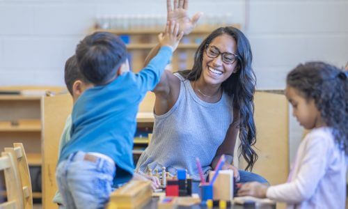 A female Montessori teacher sits with a small group of students as she works through a math lesson. They are each dressed casually and listening attentively as the teacher holds up the numbers in front of her.