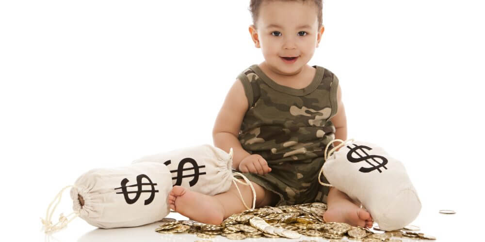 Adorable mixed race baby boy playing with a pile of plastic gold coins and canvas money bags. Isolated on white.