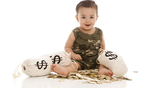 Adorable mixed race baby boy playing with a pile of plastic gold coins and canvas money bags. Isolated on white.